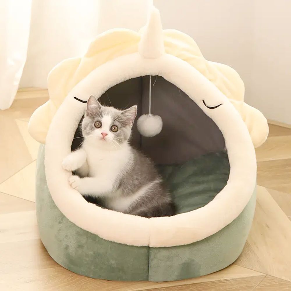 Cat lying inside a green and white pet bed with a plush ball hanging from the top.