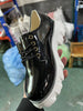 Black patent leather shoe with white sole held in hand against a blurred indoor background