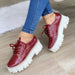 Red patent leather shoes with white soles worn with blue jeans on a light wooden floor.