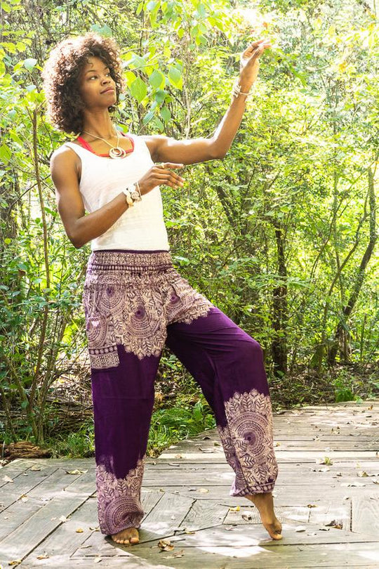 Woman in a white top and Bohemian - style purple yoga pants with floral patterns standing on a wooden path in a forest.